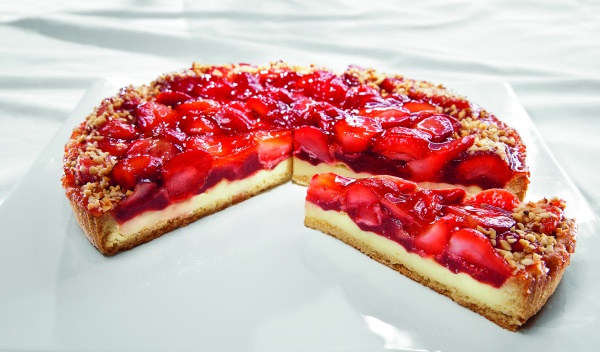 Knuspriger Buttermürbeteig mit feiner Pudding-Joghurt- Creme und einer Erdbeer-Frucht-Zubereitung gefüllt. Halbe Erdbeeren sind mit Gelee abgeglänzt und mit Haselnüssen garniert.